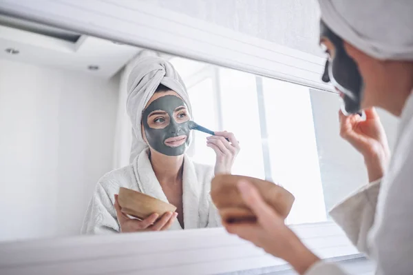 Kosmetologia, pielęgnacja skóry, pielęgnacja twarzy, spa, naturalne piękno. Piękny uśmiechnięta kobieta w domu w szlafroku z ręcznikiem stosowania maski gliny twarzy przed trądzikiem odmłodzić problem skóry — Zdjęcie stockowe