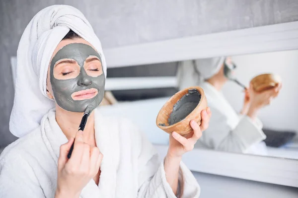 Cosmetology, skin care, face treatment, spa, natural beauty concept. Beautiful smiling woman at home in bathrobe with a towel applying face clay mask against acne to rejuvenate problem skin