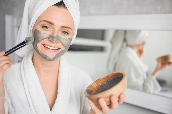 Cosmetology, skin care, face treatment, spa, natural beauty concept. Beautiful smiling woman at home in bathrobe with a towel applying face clay mask against acne to rejuvenate problem skin