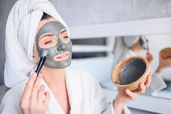 Cosmetology, skin care, face treatment, spa, natural beauty concept. Beautiful smiling woman at home in bathrobe with a towel applying face clay mask against acne to rejuvenate problem skin