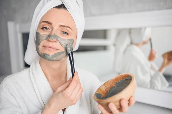 Kosmetologia, pielęgnacja skóry, pielęgnacja twarzy, spa, naturalne piękno. Piękny uśmiechnięta kobieta w domu w szlafroku z ręcznikiem stosowania maski gliny twarzy przed trądzikiem odmłodzić problem skóry — Zdjęcie stockowe