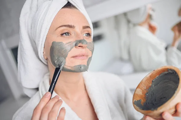 Cosmetology, skin care, face treatment, spa, natural beauty concept. Beautiful smiling woman at home in bathrobe with a towel applying face clay mask against acne to rejuvenate problem skin