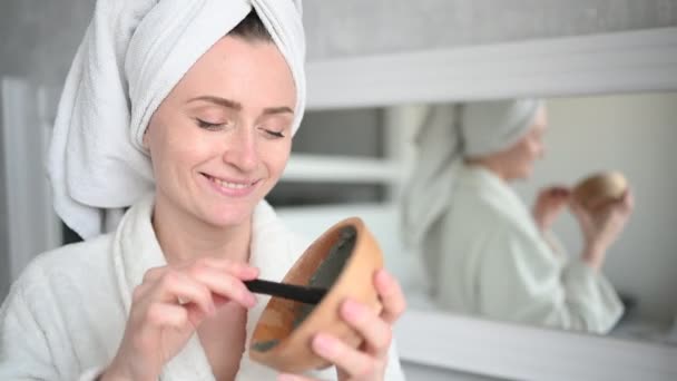 Cosmetología, cuidado de la piel, tratamiento facial, spa, concepto de belleza natural. Hermosa mujer sonriente en casa en albornoz con una toalla que aplica mascarilla de arcilla facial contra el acné para rejuvenecer la piel problemática — Vídeo de stock