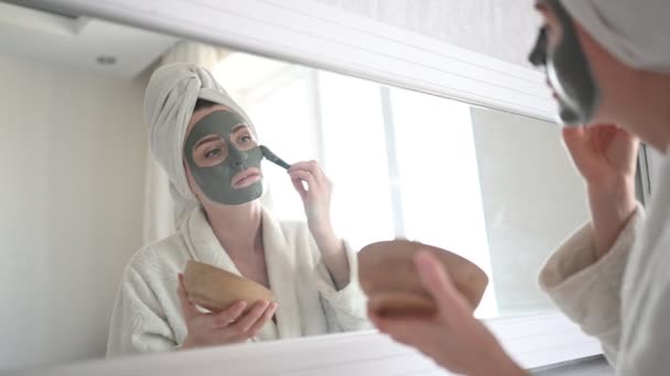 Cosmetologie, huidverzorging, gezichtsbehandeling, spa, natuurlijke schoonheid concept. Mooie glimlachende vrouw thuis in badjas met een handdoek aanbrengen gezicht klei masker tegen acne te verjongen probleem huid — Stockvideo