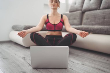 Spor kıyafetleri içinde yoga egzersizleri yapan, dizüstü bilgisayarda videolar izleyen karantina ve kapalı spor salonları boyunca oturma odasında eğitim gören formda genç bir kadın. Formda ve sağlıklı kalmak.