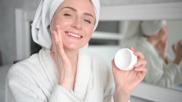 スキンケア 顔の治療 自然の美しさの概念 白い瓶から顔の保湿クリームを適用タオルでバスローブで自宅で美しい笑顔の女性 美人ルーチン — ストック動画