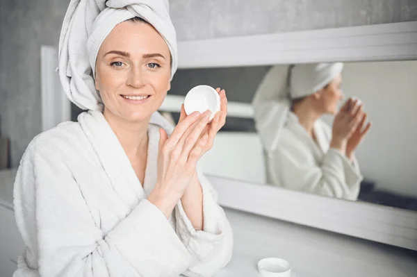 Cosmetology, skin care, face treatment, spa, natural beauty concept. Beautiful smiling woman at home in bathrobe with a towel applying face moisturizing cream from white jar. Beauty routine