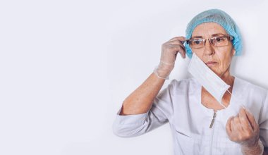 Yaşlı, üzgün, olgun bir kadın doktor ya da hemşire beyaz bir sağlık önlüğü ve eldiven giyer, yüz maskesi takar ve koruyucu ekipmanlar takar. Sağlık ve tıp konsepti. Covid-19 salgın krizi
