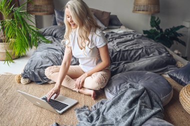 Mutlu, sarışın bir kadın çalışıyor, video görüşmesi yapıyor, rahat yatak odasındaki modern iç mekanda dizüstü bilgisayarda film izliyor. Covid-19 pandemik Corona virüsünü karantinaya alın. Ev kavramından uzak çalışma.