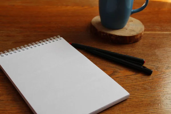 Çalışma yerinde bir defter ya da defter masanın üzerinde beyaz bir arka plan ya da ahşap bir yüzey üzerinde iki kalem, bir kalem ve bir fincan mavi çay ya da bir kahve ile ahşap bir standın üzerinde bulunur.