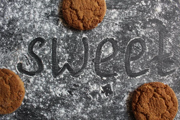 Tatlı kelimesi koyu arkaplanda yazılıdır. Yanında toz halinde veya un kaplı bir yüzey üzerinde gri bir taş. Metin için yeri olan şeker ya da kurabiye parçacıkları.
