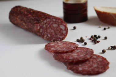 Çiğ sosis ve sosis dilimleme çubuğu beyaz bir masanın üzerinde karabiber ve biberliğin yanında duruyor.