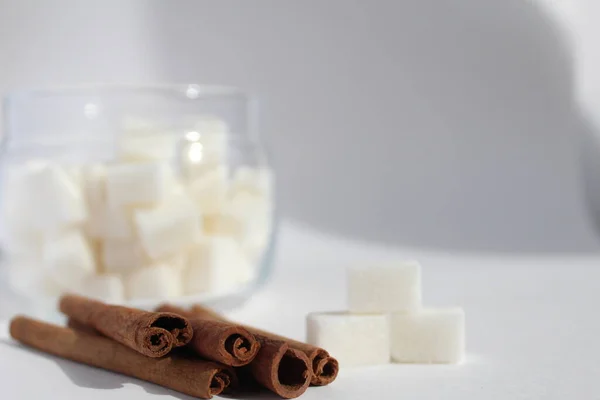 Masada üç küp şeker ve beyaz arka planda bir kavanoz şekerin yanında beş ya da daha fazla tarçın kamışı bulunmaktadır.