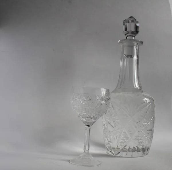 stock image crystal glass patterned pitcher decanter with vodka and a glass on a thin leg with vodka on a white gray background