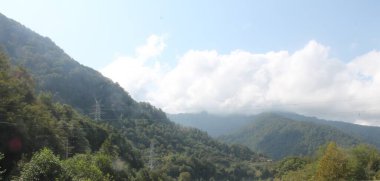 Dağların tabanı nehir boyunca yeşildir ve üzerlerinde beyaz kıvrımlı bulutlarla mavi bir gökyüzü vardır.