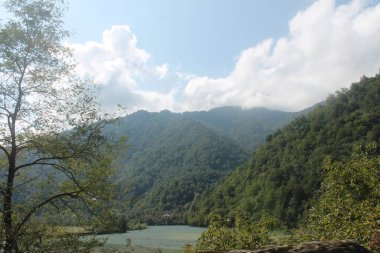 Dağların tabanı nehir boyunca yeşildir ve üzerlerinde beyaz kıvrımlı bulutlarla mavi bir gökyüzü vardır.