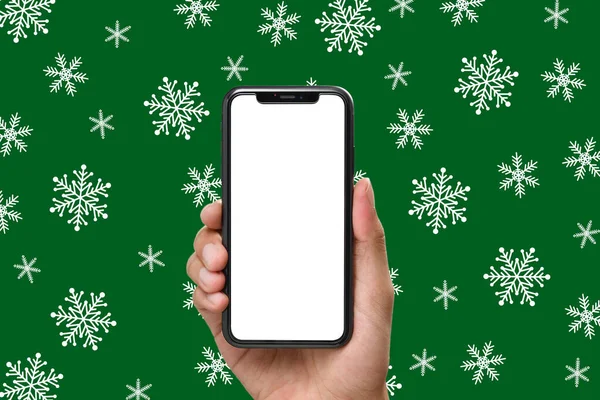 Hand holding the black smartphone with a blank screen and modern frameless design on Christmas background of snowflakes in green colour