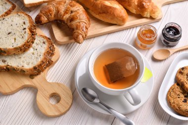 Beyaz ahşap masada, ekmekli, çörekli, kruvasan ve fırınlı çay fincanı manzarası.