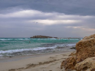 İnsanlar bulutlu bir günde deniz dalgalarında yüzerler. Kıbrıs Rum Kesimi 'nin Ayia Napa kentinde sorunlu siyanür suyu, küçük kayalık ada ve kara bulutlu gökyüzü ile pitoresk Nissi plajı. Güzel deniz tatilleri..
