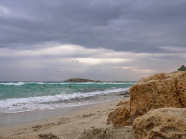 İnsanlar bulutlu bir günde deniz dalgalarında yüzerler. Kıbrıs Rum Kesimi 'nin Ayia Napa kentinde sorunlu siyanür suyu, küçük kayalık ada ve kara bulutlu gökyüzü ile pitoresk Nissi plajı. Güzel deniz tatilleri..