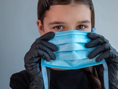 Siyah lateks eldivenli genç bir kız elinde mavi koruyucu bir tıbbi maske tutuyor. Gözler kapalı. Koronavirüs pandemisi sırasında antiviral koruma.
