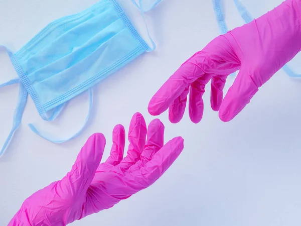 Two hands in pink latex gloves reaching out for each other. Blue protective medical face mask on a white background. Antiviral protection during coronavirus pandemia. Space for text.