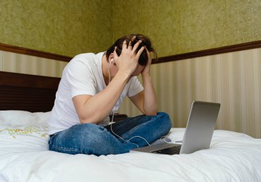 Laptop bilgisayar ve akıllı telefonla çalışan genç adam yatakta oturuyor. 