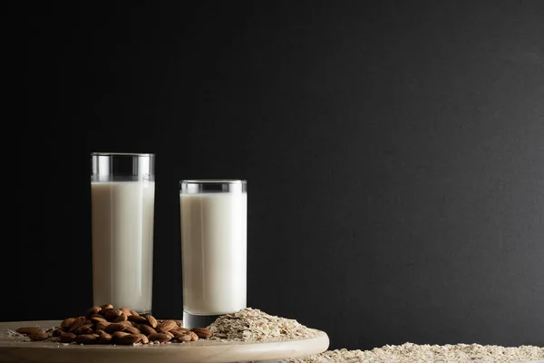 Dos vasos de leche de avena y leche de almendras con copos de avena y nueces de almendras en disco de madera. Copiar espacio, fondo negro . — Foto de Stock