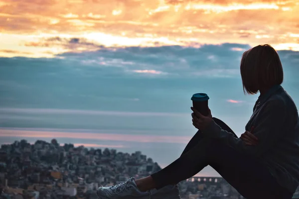 街の上に夕日の空を見て丘の上に座ってコーヒーのカップを持つ女性 — ストック写真