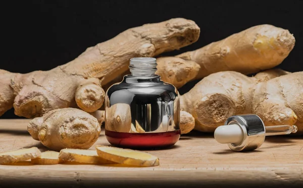 Bottle of cosmetics with glass pipette, ginger oil extract on wooden board with ginger roots. Black background, copy space. — Stock Photo, Image