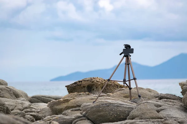 Trípode con teléfono móvil conectado instalado en las rocas por el mar de vídeo lapso de tiempo de disparo en el teléfono inteligente. Concepto de creación de video y fotografía. Copiar espacio . — Foto de Stock