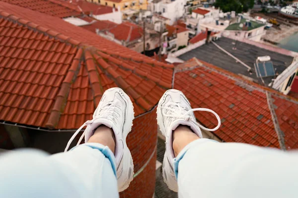 Woman sitting on the edge of building and her legs with white sneakers dangling against red rooftops