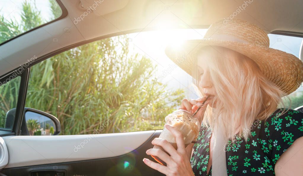 Attractive blonde woman with straw hat drinking coffee in a car during road trip.