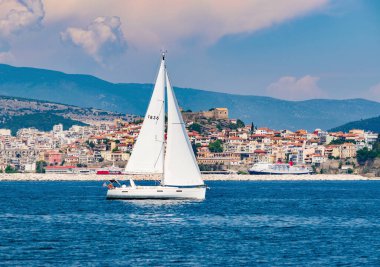 Mavi denizde yüzen beyaz yelkenli teknenin arkasında güzel küçük bir şehir var.