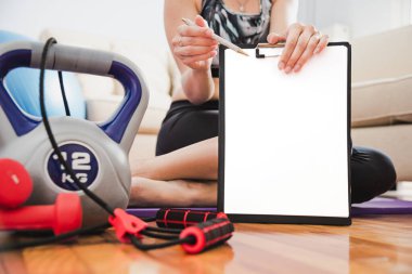 Atletik kadın boş bir a4 kâğıt maketi tutuyor. Spor aletleri oturma odasında. Ev ödevi rutin konsepti. Boşluğu kopyala.