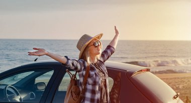 Hasır şapkalı genç kadın arabanın yanında duruyor ve günbatımının tadını çıkarıyor..