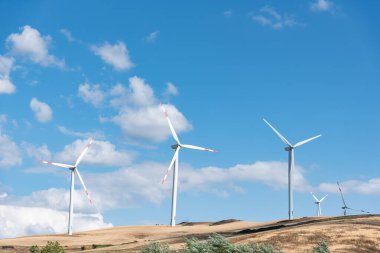 Tarlada mavi gökyüzü ve beyaz bulutlu üç rüzgar türbini var..