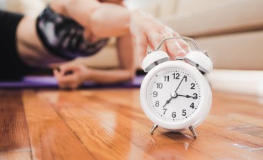 White classic alarm clock on wooden floor in the living room. Unfocused woman shuts alarm clock during 10 minutes home workout. clipart