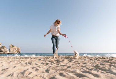 Genç sarışın kadın sahilde köpekle yürürken eğleniyor.