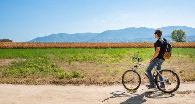 Bisiklet sürerken dinlenen genç adam dışarıda manzaranın keyfini çıkarıyor.