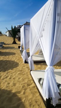 Sahilde beyaz perdeleri ve büyük yatağı olan ahşap şezlonglar. Plaj konseptinde lüks yaz tatili.