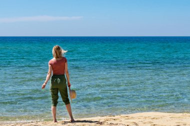 Hasır şapkalı genç bir kadın sahilde yalnız başına yürüyor. Yaz tatillerinin tadını çıkarıyorum. Boşluğu kopyala.