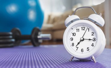 Alarm saatinin düşük açılı görüntüsü. Matt 'in evde olması. Arka planda odaklanmamış kanepe, fitness topu ve dambıllar var. Egzersiz yapma zamanı.