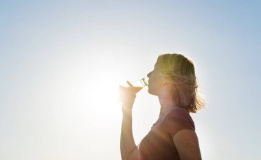 Genç çekici kadın taze portakal suyu içiyor ve gün batımında parlak güneş ışığının tadını çıkarıyor. Sağlıklı yaşam tarzı ve eğlence anlayışı.