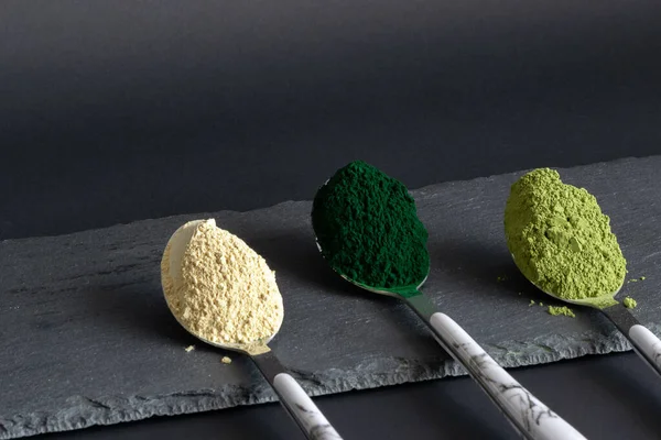 Löffel mit Spirulina, Matcha und Macapulver auf schwarzem Steinbrett. Abgewinkeltes Bild. Konzept für Superfood-Pulver. — Stockfoto
