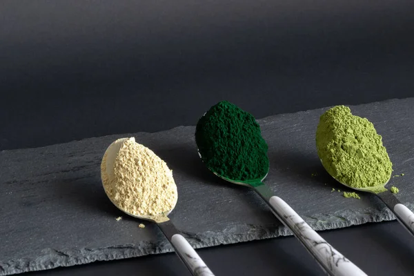 Cucchiai con polvere di spirulina, matcha e maca su tavola di pietra nera. Immagine angolata. Concetto di polveri superalimentari . — Foto Stock