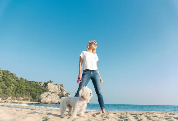 年轻的金发女人和狗在海滩边散步很开心 — 图库照片