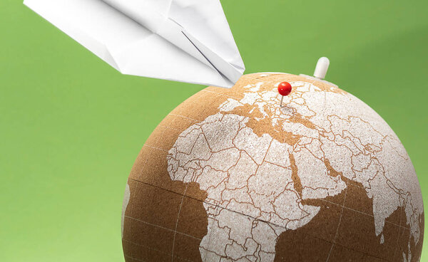 Cork globe with red pin on European region and paper plane landing. Green background. Holiday traveling concept.