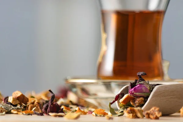 Ervas de chá secas misturadas e flores em pá de especiarias de madeira com chá sem foco em vidro turco no fundo . — Fotografia de Stock
