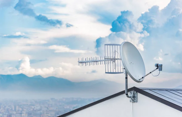 Evin çatısına yerleştirilmiş uydu anteni ve televizyon anteni. Arka planda bulutlu bir gökyüzü var..